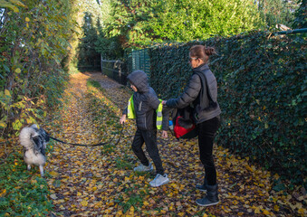 Mother and son walking with their Keeshond dog along a sunlit autumn path covered with fallen yellow leaves, child wearing a reflective vest for safety during daily outdoor activity