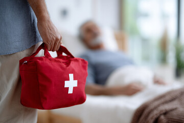 Caregiver with red medical bag assisting sick elderly man in bed at home or nursing facility