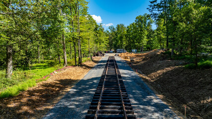 Construction crews are laying down railway tracks through a lush, green forest. The area is cleared, and heavy machinery is visible nearby under a bright blue sky with scattered clouds.