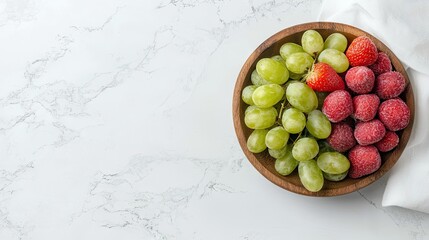 Overhead shot of a wooden bowl filled with green grapes, strawberries, and raspberries, set on a white marble surface. A white cloth is partially visible.