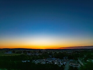 A vibrant sunset casts warm colors across the sky above a small town. Fields stretch out in the distance, creating a serene atmosphere. The peaceful scene shows nature’s beauty as day turns to night.