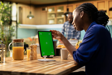 Vertical green screen on tablet with young black woman enjoying breakfast at the table while partner prepares the meal. Representing modern lifestyle, technology and togetherness.