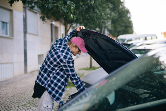 Young man diagnosing car engine on residential street during daytime