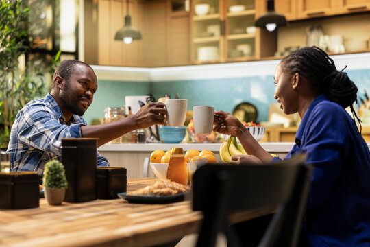 African american lovers clinking mugs with coffee in the morning, enjoying weekend bonding and sitting together. Sweet affectionate man and woman chatting at the breakfast table.