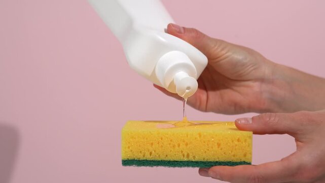 Close-up of female hands pouring dishwashing gel onto yellow sudsy sponge on pink isolated background. Concept of kitchen routine, maintaining cleanliness, shine, and freshness on dishes.