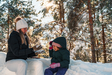 Mother and daughter sitting together on a snowy forest floor, enjoying a sunny winter day, bundled in warm clothes, playing joyfully with snowflakes and creating happy memories