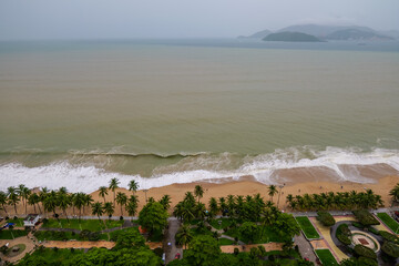Coastal view of a cloudy day beach with palm trees, ocean waves, and distant mountains in a resort city.