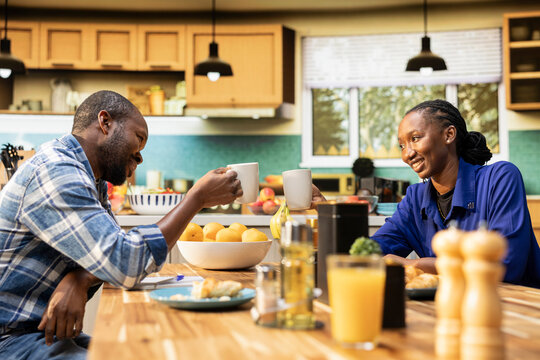 Black boyfriend and girlfriend saying cheers over cups of coffee, clinking mugs and enjoying slow morning on the weekend. African american people sharing a romantic moment at home. - Powered by Adobe