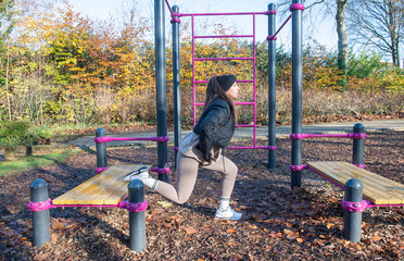Woman exercising outdoors doing forward lunge on fitness equipment in a public park, focusing on leg workout and healthy active lifestyle during autumn season