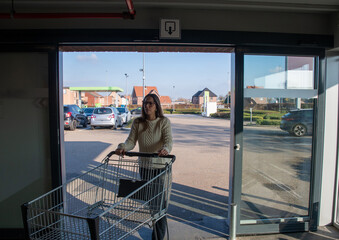 Woman standing with an empty shopping cart, pushing it from inside a supermarket, moving through an automatic glass door opening towards the bright outdoor parking lot area under a clear blue sky