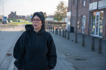 Fototapeta premium Woman standing on an urban street, wearing a casual black hoodie and glasses, looking away with a thoughtful expression while reflecting on personal thoughts or future plans in a city environment