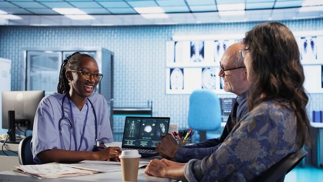 Medical assistant consulting couple of patients about test results and MRI scans, reviewing records to create the diagnostic report. Patient care management in a medical cabinet. Camera A.