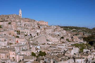 Obraz premium panoramic view of matera's sassi, an unesco world heritage site, featuring historic cave dwellings and the iconic bell tower under a bright blue sky. a testament to ancient architecture and history