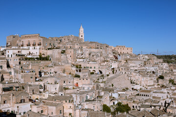 Obraz premium panoramic view of matera's sassi, an unesco world heritage site, featuring historic cave dwellings and the iconic bell tower under a bright blue sky. a testament to ancient architecture and history