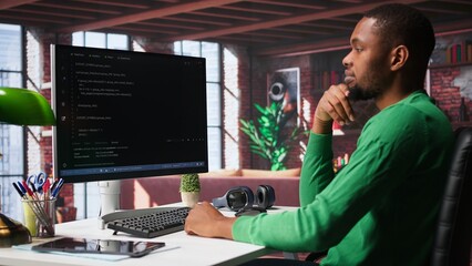 Black technician in home office looks at UI on computer monitor, typing and executing programming code. IT worker using coding application to write and test code in modern apartment. Camera B.