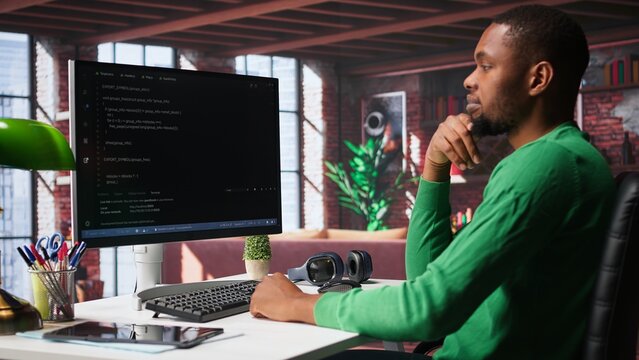 Black technician in home office looks at UI on computer monitor, typing and executing programming code. IT worker using coding application to write and test code in modern apartment. Camera B. - Powered by Adobe