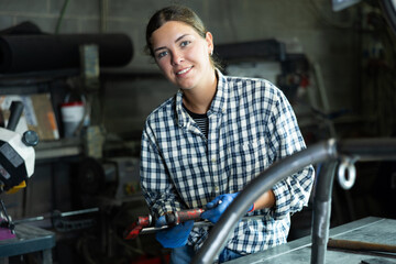 Young girl, professional technician with clamp in hands, wearing checkered shirt and gloves, posing in industrial metal workshop, smiling confidently and friendly at camera..