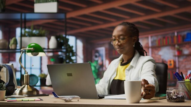 Remote employee drinking coffee at home while looking at social media posts on notebook. African american woman enjoying hot beverage and reading ebooks on laptop, camera A