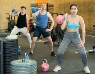 Young brunette fitness woman working out her biceps and shoulders with kettlebell during CrossFit class in gym. Functional training concept