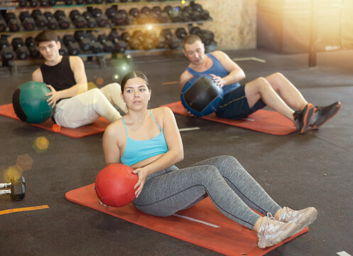 Sporty young girl doing twisting sit-ups with medicine ball to strengthen abs muscles during group training in gym. Active lifestyle concept