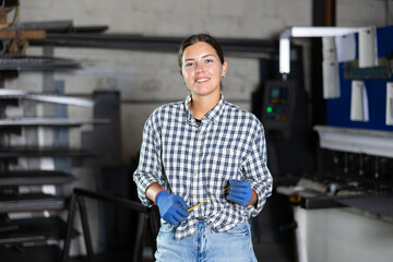 Smiling young female worker in casual plaid shirt, jeans and gloves posing at workplace in metalworking shop, surrounded by tools and machines, holding tape measure, ready to mark workpieces..
