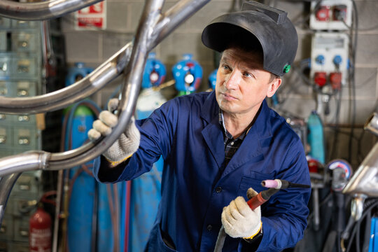 Focused experienced male welder in blue workwear, protective gloves and helmet welding structure of metal pipes in workshop, holding torch and carefully inspecting quality of weld seam - Powered by Adobe