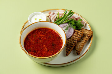 A rich, red soup sits in a bowl beside crunchy bread, crisp greens, and onions, creating a colorful display of flavors for a delightful dining experience