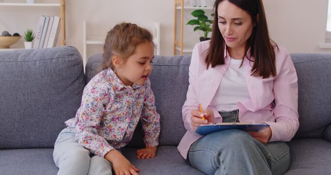 Young female counselor and behavior correction talking to little girl during therapy session. Psychologist taking notes on clipboard while sitting on sofa in office with child. Child therapist concept