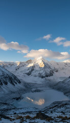 Snow covered mountain peak reflecting in frozen alpine lake under blue sky with soft clouds at sunrise, serene cold landscape with glacier and rocky foreground