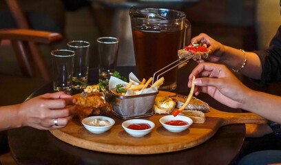 A lifestyle food scene featuring hands reaching for fries, bruschetta, and fried appetizers served on a wooden board. Warm restaurant lighting creates a cozy social dining atmosphere.