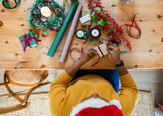 Gift wrapping scene unfolding on light wooden table with evergreen wreath, red ribbon, and berries