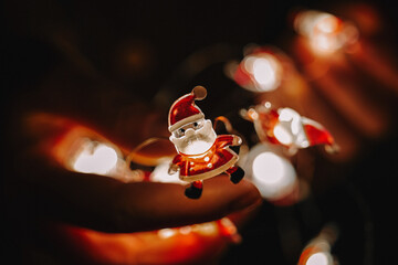 Hands Framing Miniature Santa Light, Warm Glow, Lifestyle Blogger Gently Holding Ornament For Candid Shot, Sparkling Bokeh And Soft Amber Tones, Creative Evening Ritual Focused On Festive