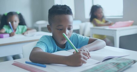 Obraz premium Writing boy wearing light blue T-shirt focusing on notebook at classroom, holding green pencil
