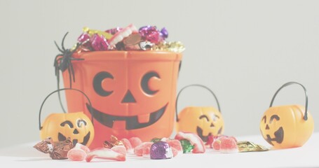 Fototapeta premium Showing large orange pumpkin bucket holding candy on white table, three small pumpkin pails, spider