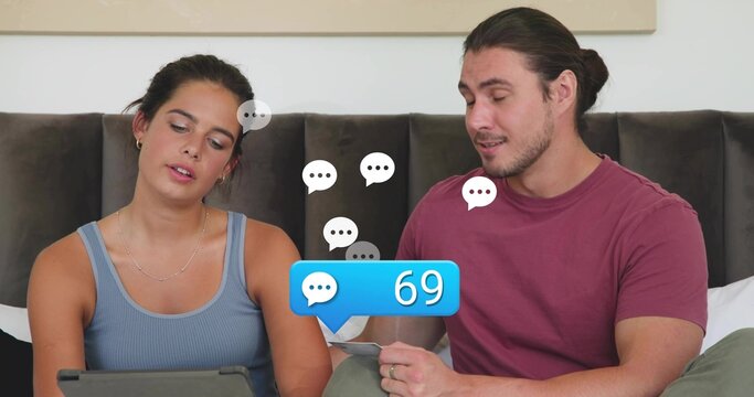 Browsing couple in blue-gray tank and maroon tee on bed holding tablet, paper, 69 message bubble