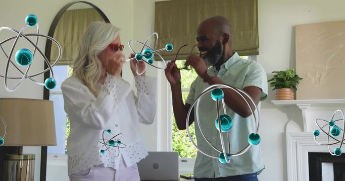 Smiling senior couple interacting with floating atom models at home, with laptop and red sunglasses