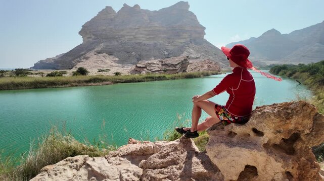 Woman sits on a rock at Wadi Sinaq in Oman, gazing over turquoise oasis water and rugged canyons, soaking in serene desert landscape and peaceful outdoor solitude during travel