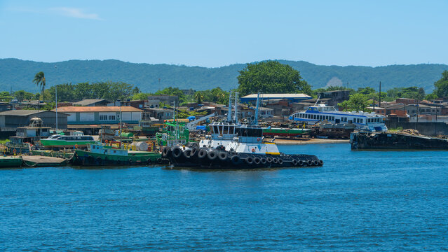 Harbor scene with tugboats and barges docked near a coastal town with green mountains
