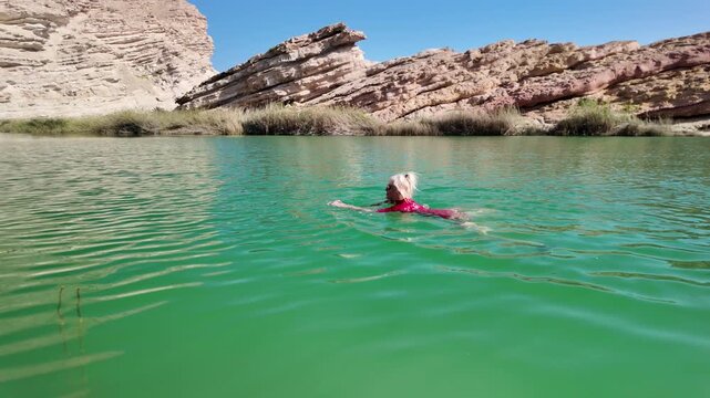 Woman enjoying a refreshing swim in the turquoise waters of a natural Wadi Sinaq pool in Oman, surrounded by desert mountains and lush vegetation, finding relaxation and leisure during travel