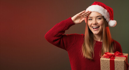 Smiling young woman in Santa hat saluting and holding a Christmas gift box. Cheerful holiday portrait on red and green background. Copy space for text