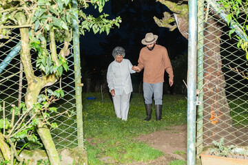 Bearded Colombian grandson guiding Afro-Colombian grandmother through garden gate at night.