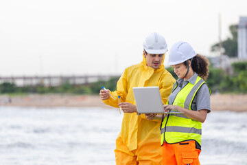 Environmental engineers collecting water samples and reviewing data on laptop, Coastal inspection team testing water quality and analyzing environmental data with safety gear and digital tools