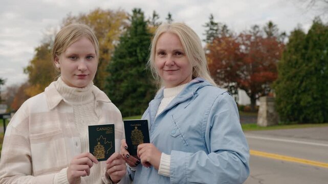 A young woman and a middle-aged woman stand outdoors and show their Canadian passports, holding the documents toward the camera.