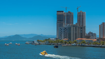 Sunny coastal cityscape with modern high-rise buildings under construction, boats sailing on blue water, and mountains in the background