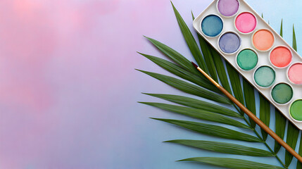 Pastel watercolor paint palette with a paintbrush resting on a tropical palm leaf.