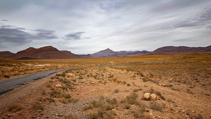 Long, winding asphalt road traversing the desolate arid background desert landscape, leading towards rugged, distant mountains under a dramatic, cloudy sky in Morocco