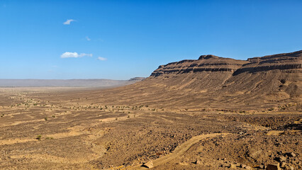Vast arid desert background landscape with tiered rock plateaus and steep cliffs, featuring a dry, winding track under a bright blue sky in Morocco