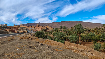 Roadside view of a traditional Moroccan village built of clay and mud bricks, nestled against a lush palm grove with an arid mountain background under a bright, cloud-streaked sky