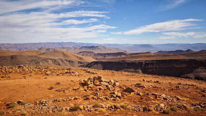 Vast rugged panorama of the arid Atlas Mountains landscape in Morocco, featuring striking red-orange rocks, deep dry valleys, and a wide blue sky with streaky clouds