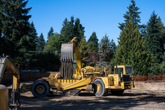 Excavator bucket full of dirt pouring it into a scraper machine, with large pit being dug as part of a large earthworks construction project on a sunny day
